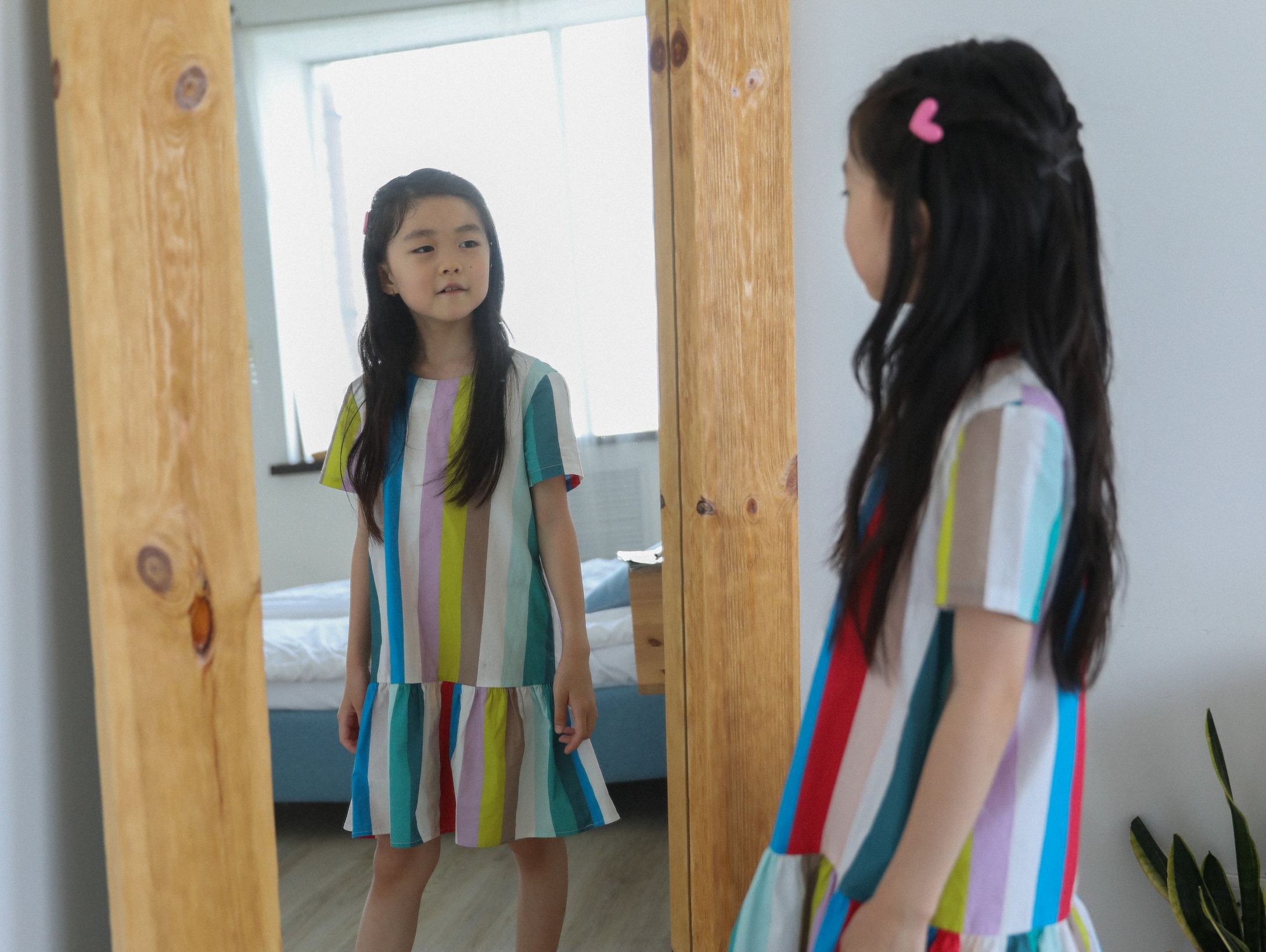 Cute Asian girl in bright dress with colorful stripes reflecting in mirror with wooden frame