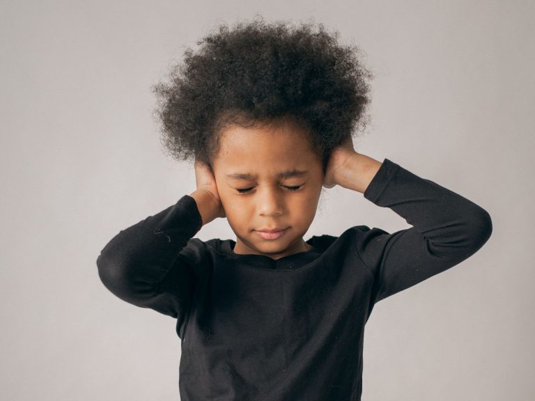 Dissatisfied African American girl in casual wear covering ears while standing on white background with closed eyes in light studio