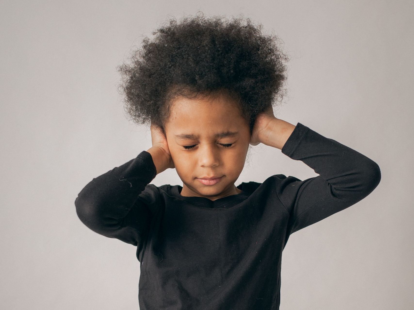 Dissatisfied African American girl in casual wear covering ears while standing on white background with closed eyes in light studio