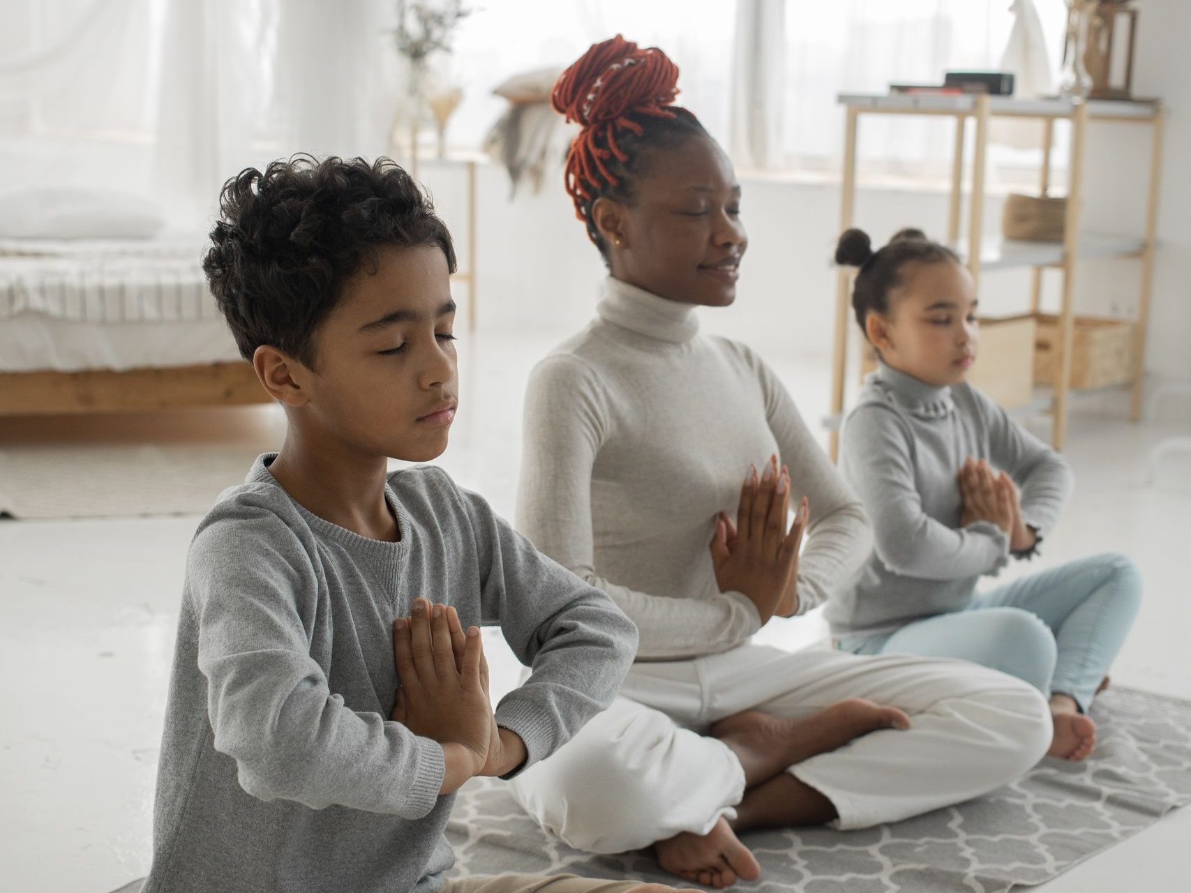Full body of happy young black mother with son and daughter sitting on rug in Lotus pose and meditating together with closed eyes and prayer hands