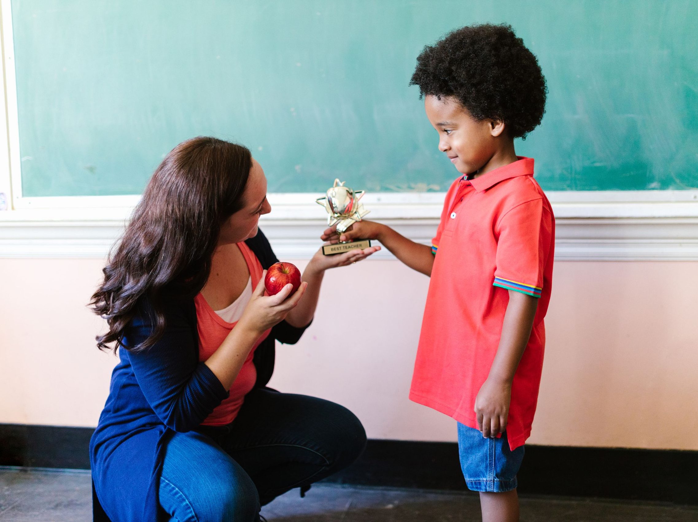 Teacher kneeling and handing a trophy to a smiling young boy in a classroom setting.