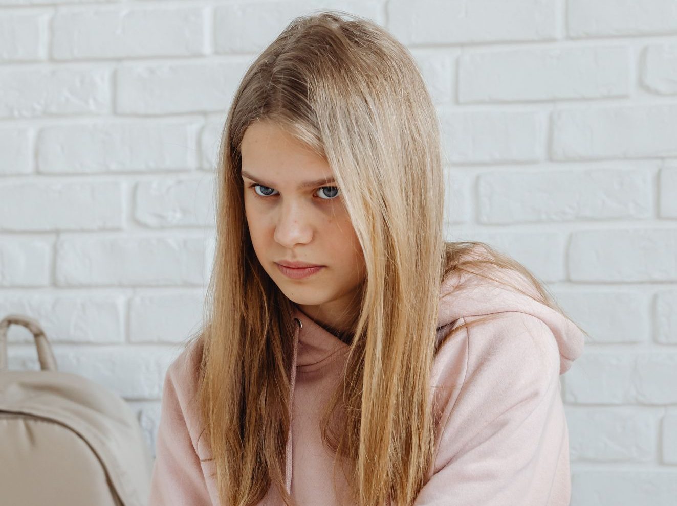 Teenage girl in pink hoodie sitting against white brick wall with a backpack.