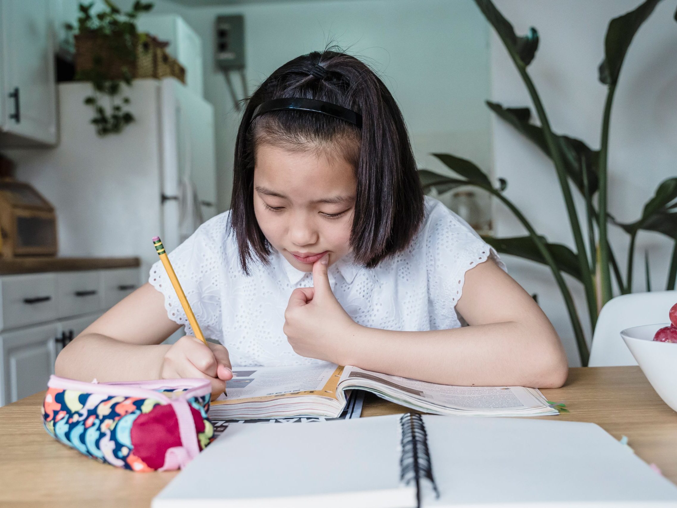 A young girl in a kitchen intently doing her homework, writing in a notebook.