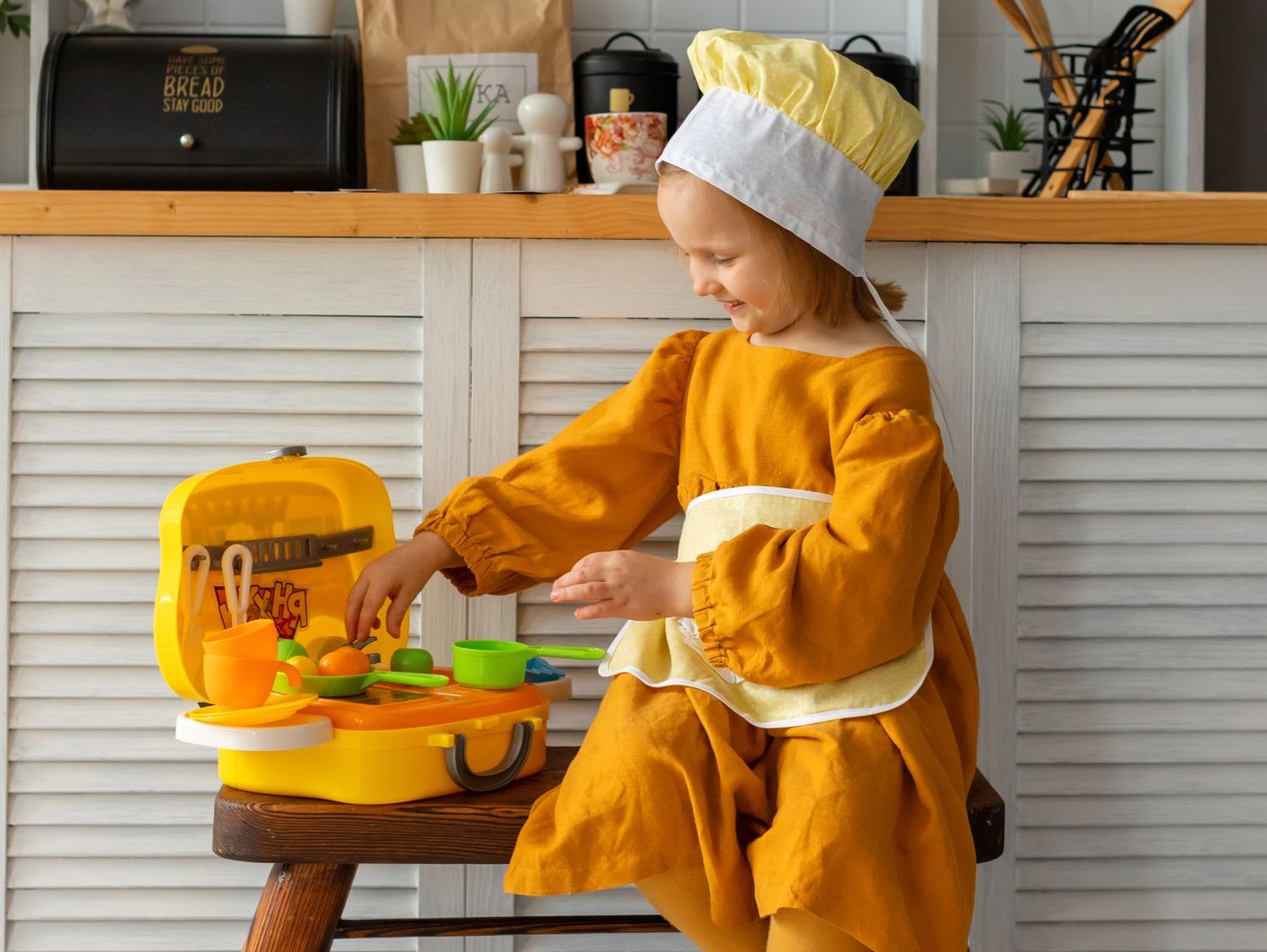A young girl wearing a chef hat plays with a toy kitchen set indoors.