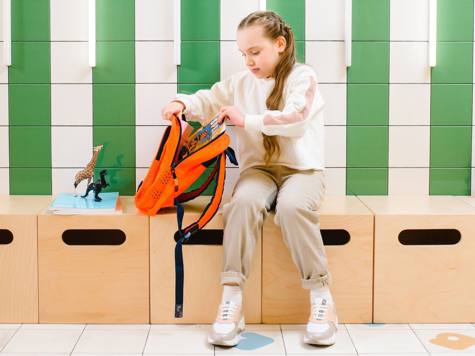 Young girl organizes backpack while sitting indoors with modern green tile backdrop.