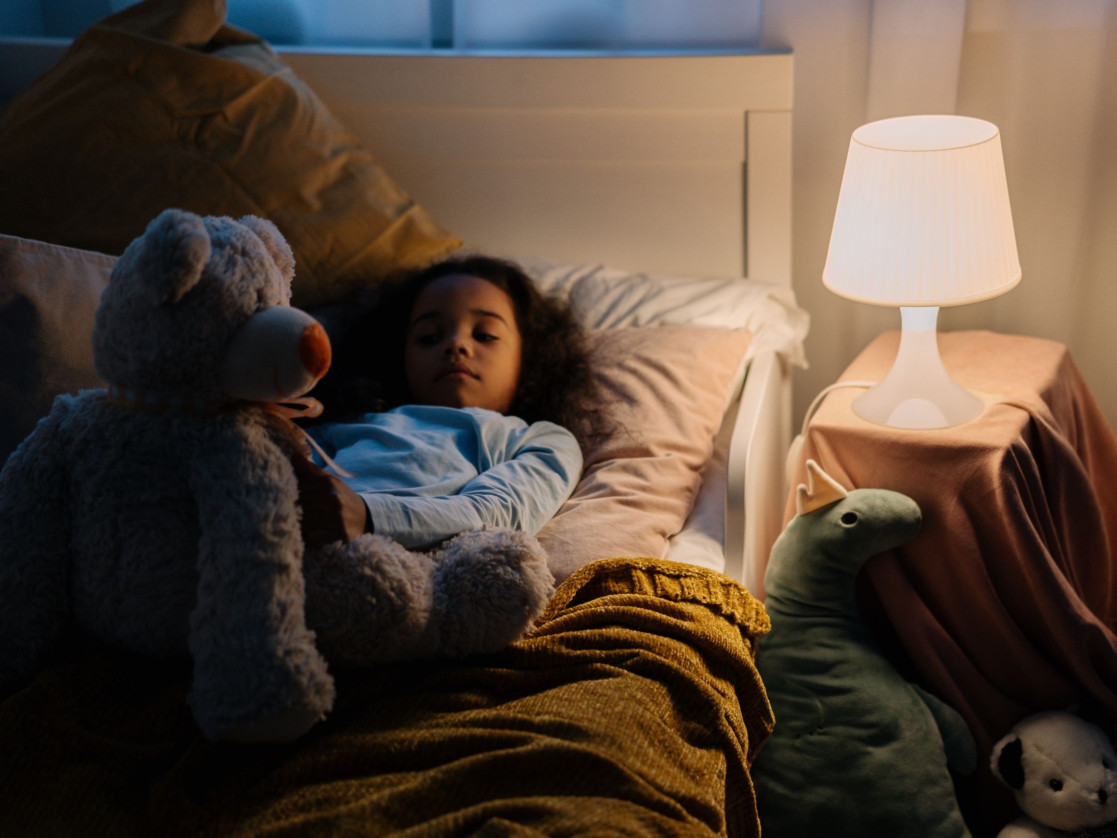 Young girl resting in bed with a teddy bear and toys, illuminated by a bedside lamp.