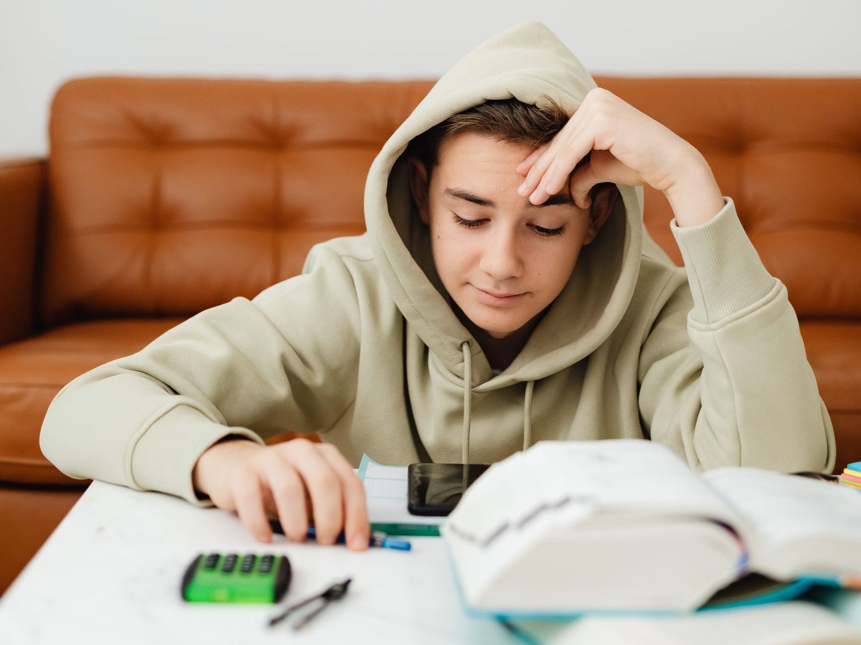 Teenager in a hoodie studying at home with books and calculator on the table.